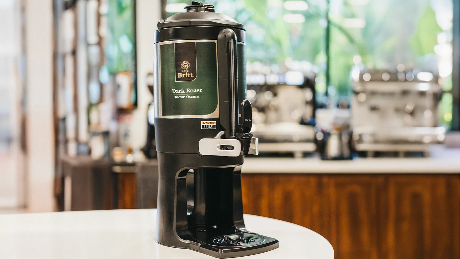 Black coffee maker with green label on a white surface, blurred cafe background