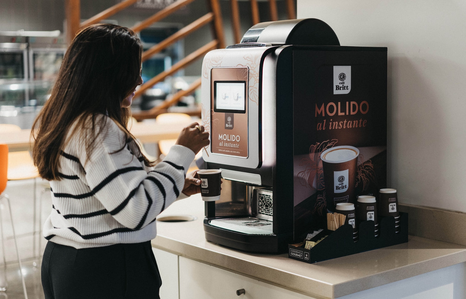 Employee using a Café Britt commercial coffee machine in a modern office break room.
