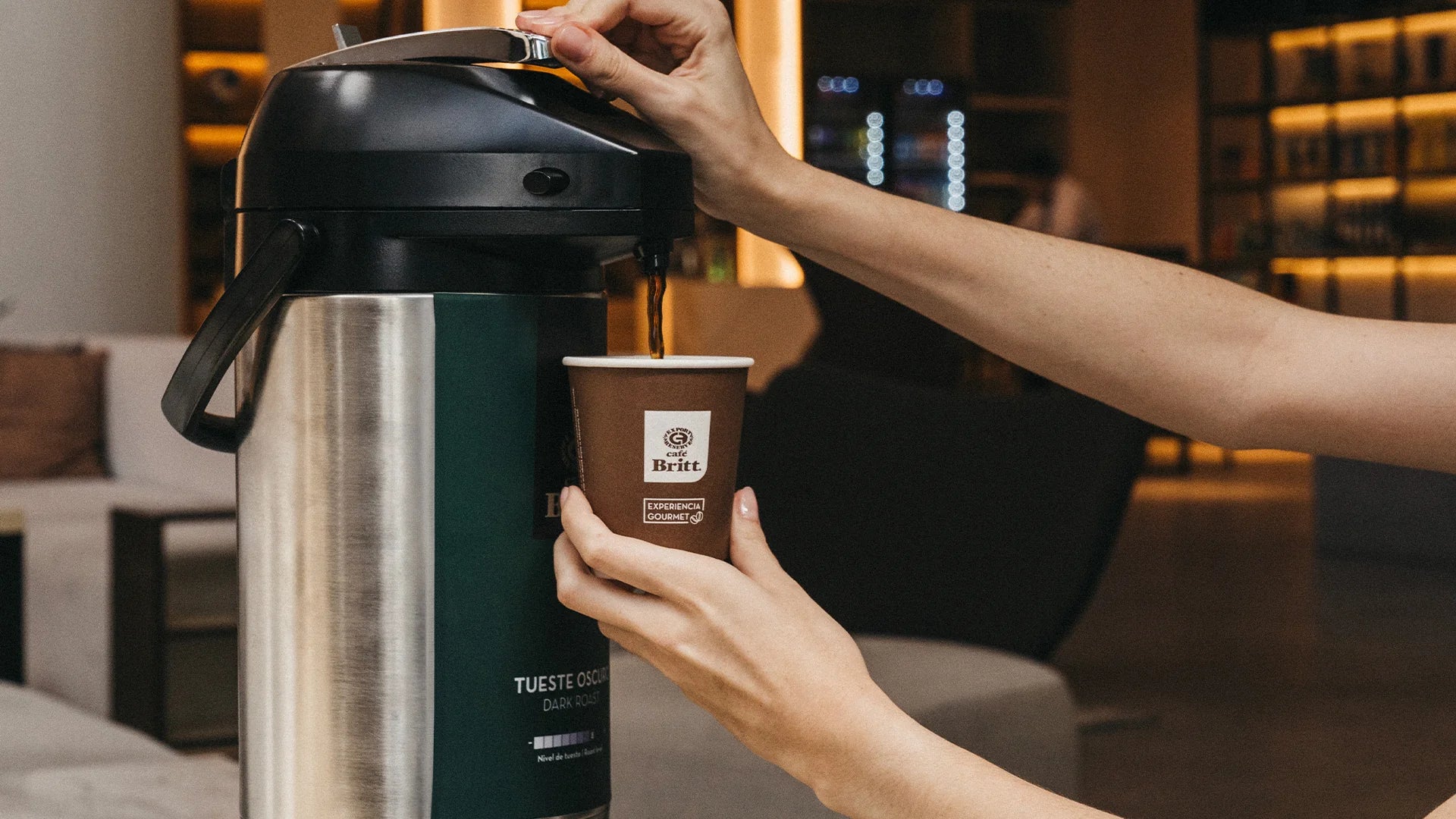Person pouring coffee from a thermos into a branded paper cup in a cozy indoor setting.