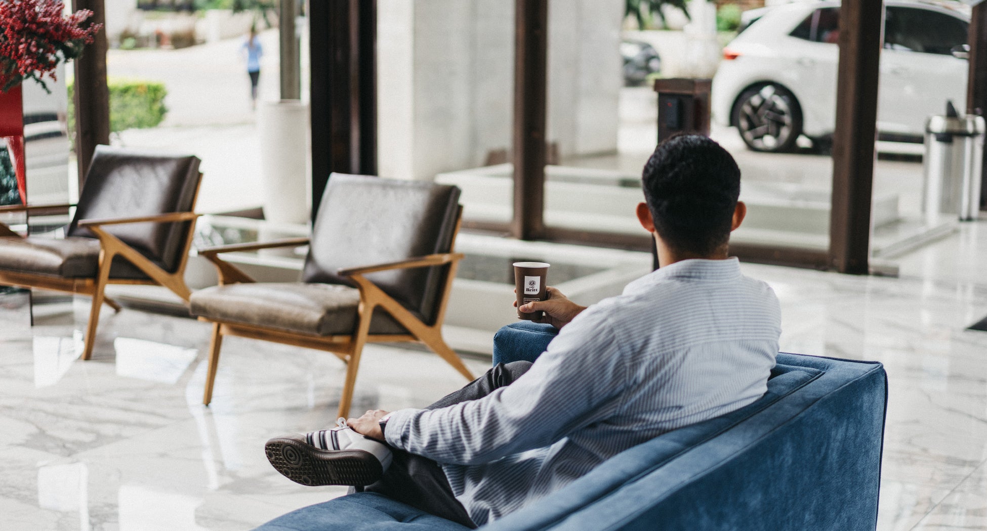 Man sitting on a blue chair in a modern cafe, holding a Britt coffee cup.