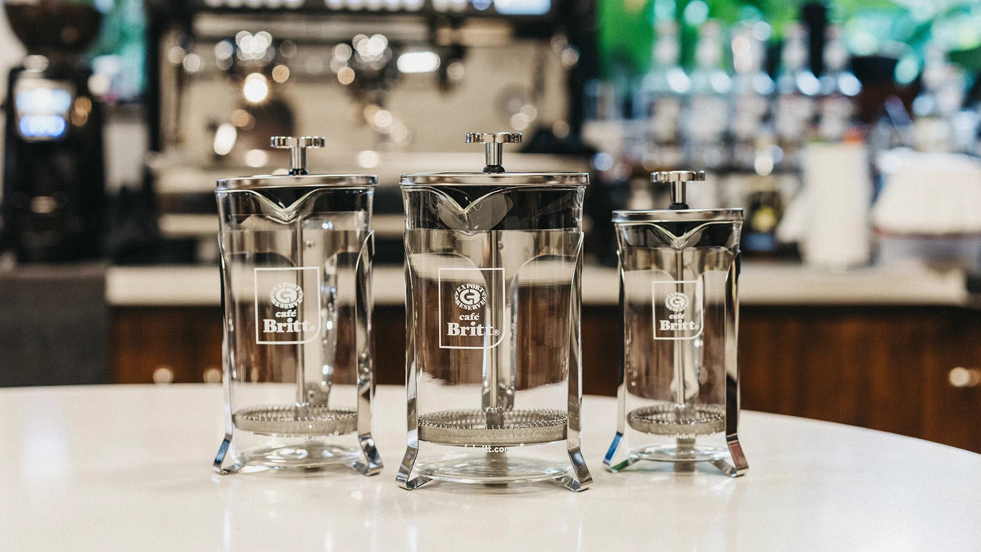 Three glass coffee presses with a brand logo on a counter in a bar setting.