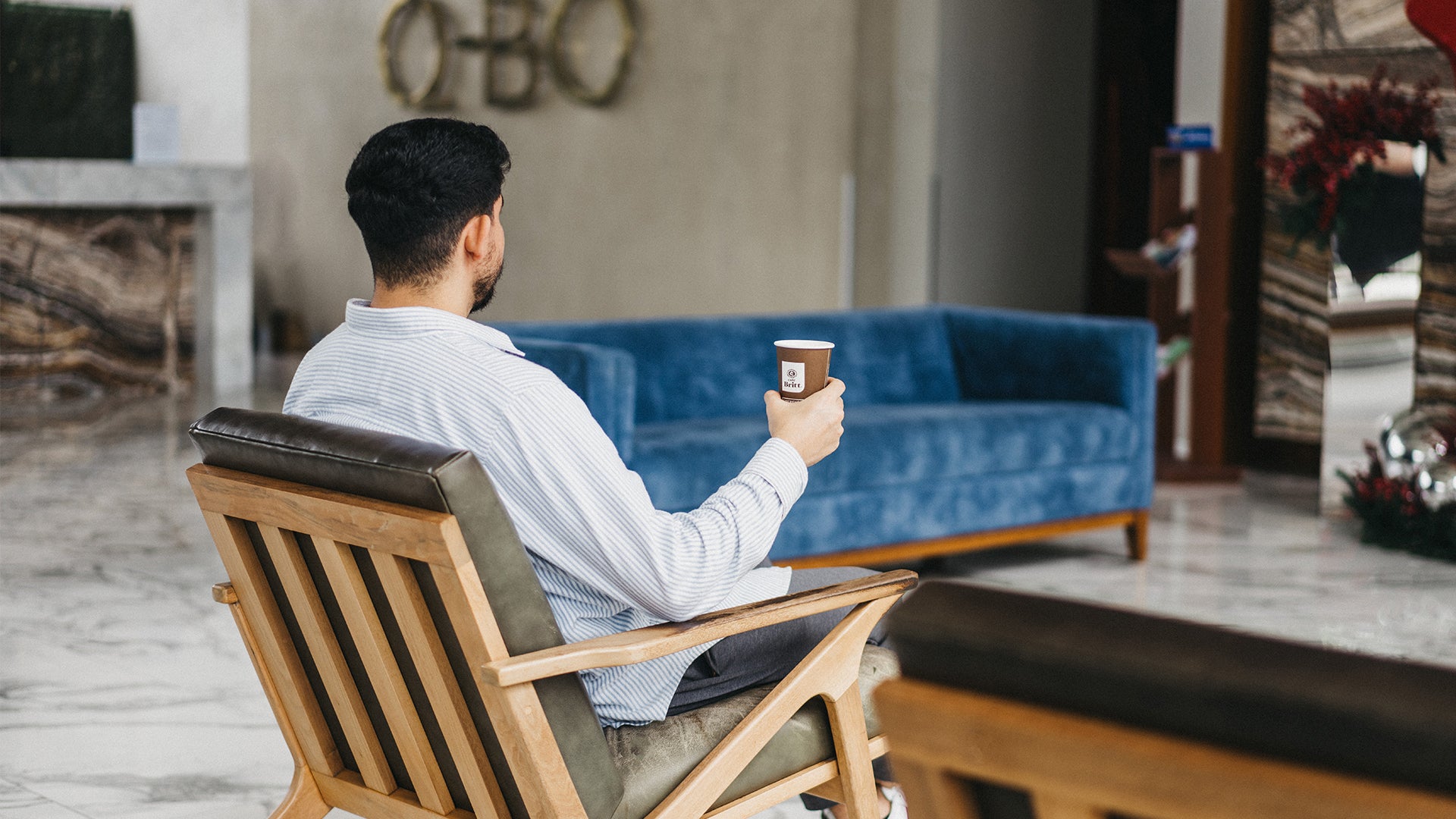 Man sitting in a chair holding a coffee cup in a room with a blue couch and decorative items.